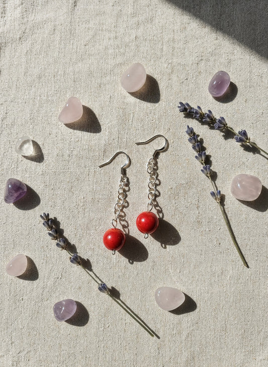 Pair of earrings with red beads on a textured surface with lavender and crystals.