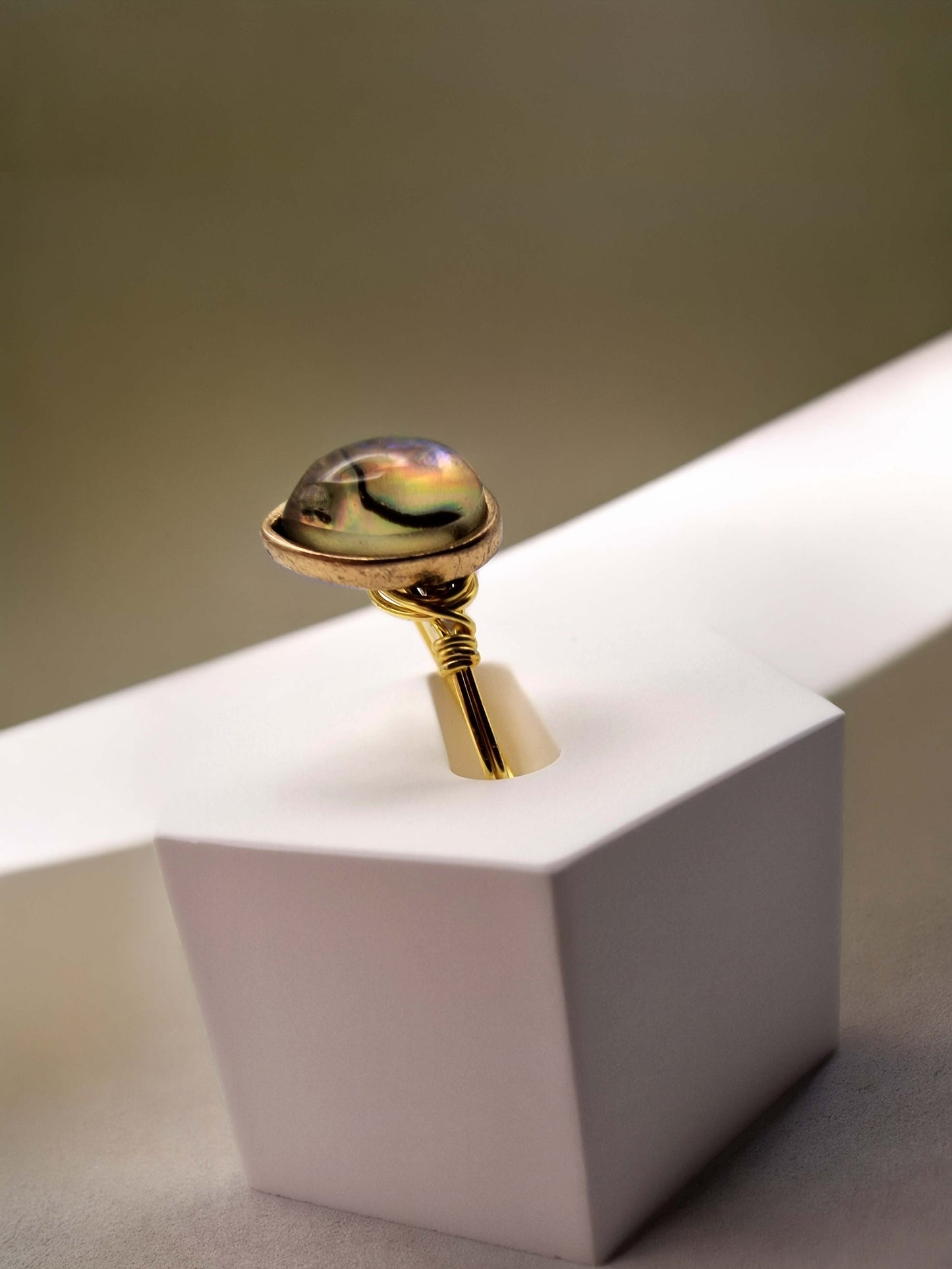 A gold ring with a polished, domed iridescent shell gemstone, displayed on a white geometric pedestal against a neutral beige background.