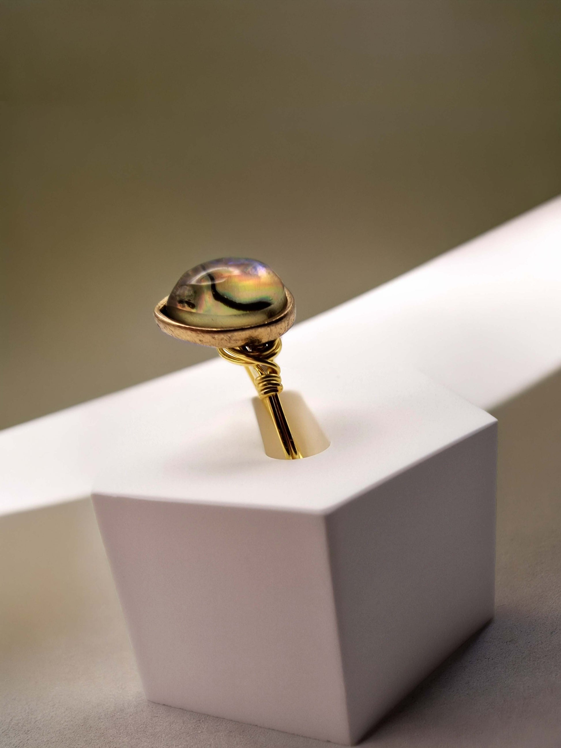 A gold ring with a polished, domed iridescent shell gemstone, displayed on a white geometric pedestal against a neutral beige background.